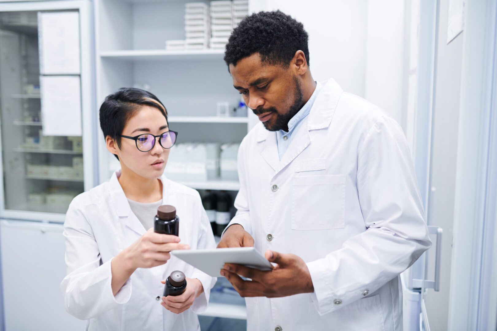 Serious thoughtful multi-ethnic pharmacists in lab coats standing in drugstore and discussing new medications while using tablet to search for information in database