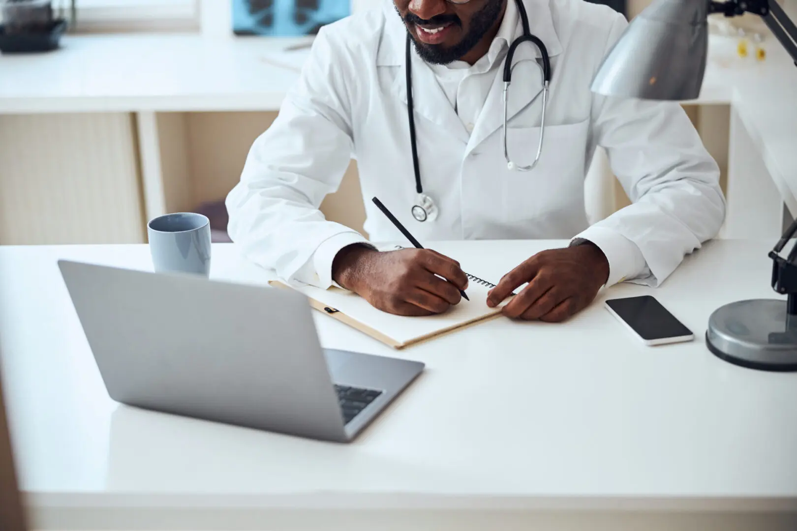 A doctor writing on paper at desk with laptop in background.